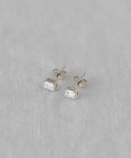 The Ivy Emerald Cut Studs feature clear gemstones set in white gold with butterfly backs, elegantly displayed on a soft, light gray fabric background.
