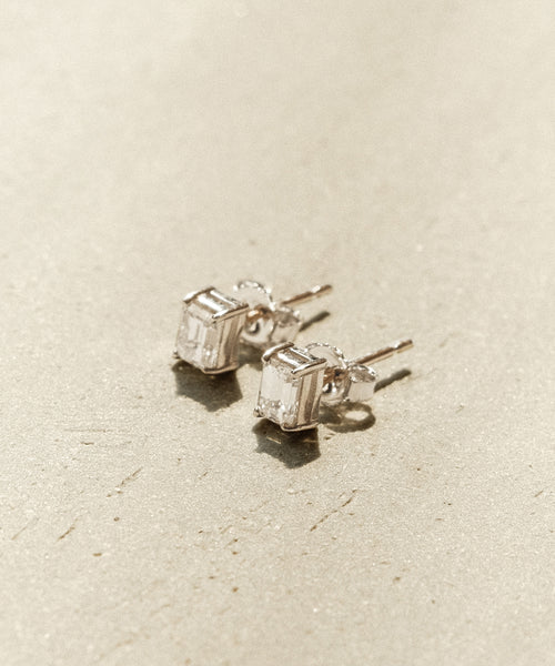 The Ivy Emerald Cut Studs feature minimalist silver settings with rectangular clear gemstones, showcased on a textured beige surface.