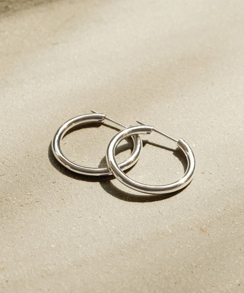 Medium Eloise Hoops in white gold are displayed on a textured beige surface, illuminated by soft natural light that casts subtle shadows.