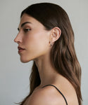 A woman with long brown hair is shown in profile against a plain background, wearing a thin-strapped top and the Medium Eloise Hoops.