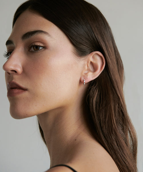 A woman with long brown hair and natural makeup looks to the side, wearing a thin black strap top and the delicate Merit Baguette Hoops earrings, against a plain light gray background.