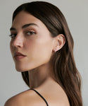A woman with long brown hair and light skin looks over her shoulder at the camera, wearing a thin black strap top and elegant Merit Baguette Hoops earrings. She has a neutral expression, set against a plain background.