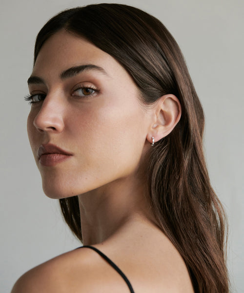 A woman with long brown hair and light skin looks over her shoulder at the camera, wearing a thin black strap top and elegant Merit Baguette Hoops earrings. She has a neutral expression, set against a plain background.