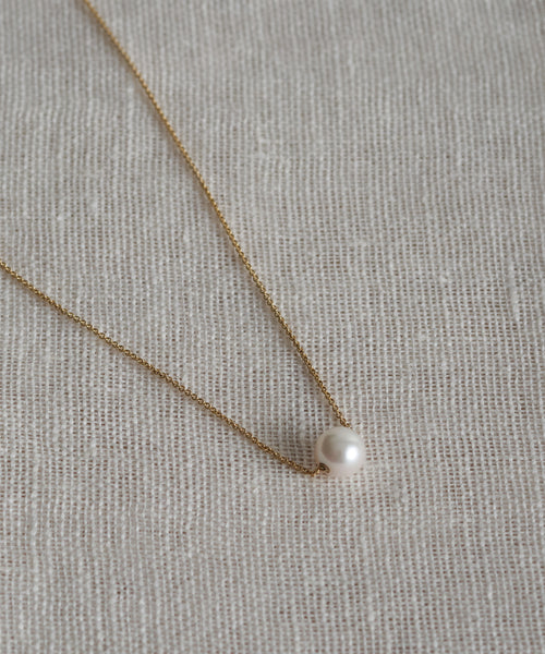 The Philo Pearl Necklace features a delicate 14k gold chain with a single white pearl pendant, showcased on textured beige fabric—a timeless fine jewelry piece.