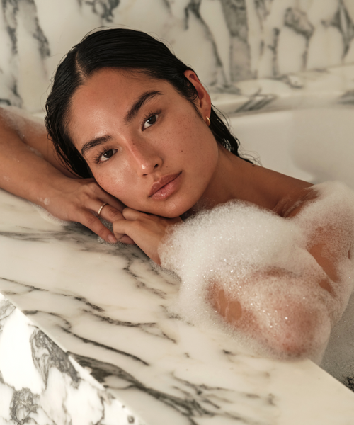 A woman with wet hair relaxes in a marble tub filled with bubbles and hydrating oil from the Relaxation Bath Set, resting her head on her arms and gazing calmly at the camera amid elegant white and gray marble surfaces.