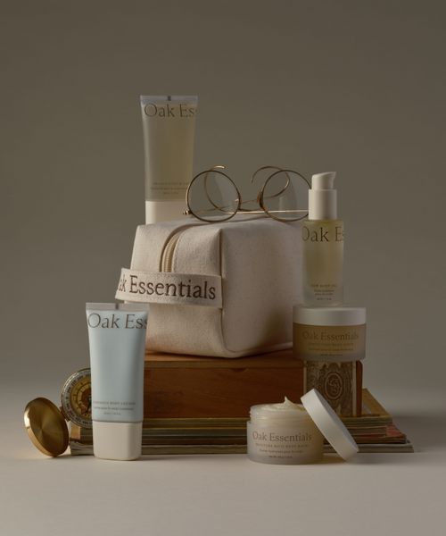 The Travel Body Routine features Oak Essentials skincare minis—TSA-approved tubes, jars, and bottles—displayed with a canvas bag, books, eyeglasses, and a compact mirror against a neutral backdrop for effortless travel skincare.