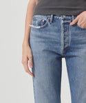 A person in AGOLDE’s 90s Jean—a blue, high-waisted denim—paired with a dark ribbed top stands with one hand in their pocket against a plain light background.