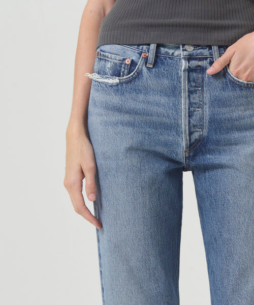A person in AGOLDE’s 90s Jean—a blue, high-waisted denim—paired with a dark ribbed top stands with one hand in their pocket against a plain light background.
