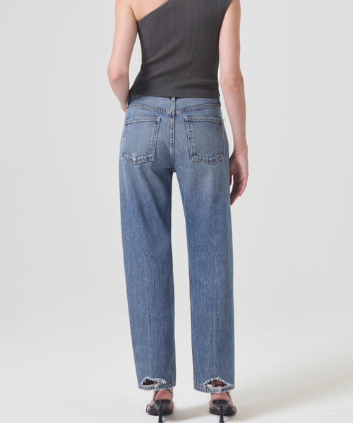 A person stands facing away, wearing high-waisted 90s Jean by AGOLDE in organic cotton with frayed hems, paired with a black one-shoulder ribbed top and black heeled sandals against a plain light background.