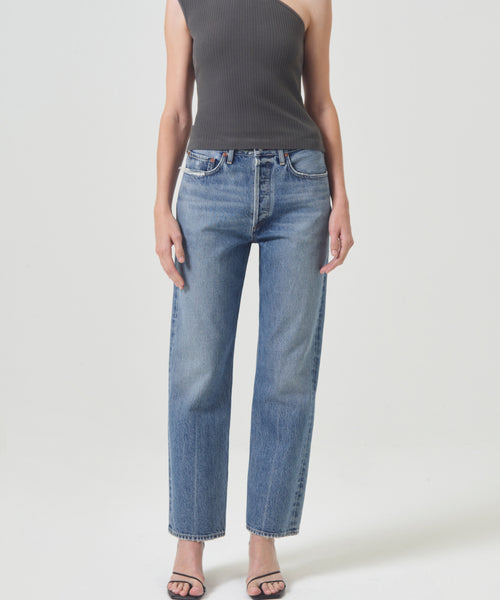 Person wearing a sleeveless, one-shoulder black top, high-waisted 90s Jean by AGOLDE crafted from organic cotton, and black heeled sandals, standing against a plain light background.