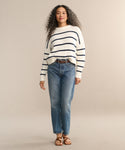 A woman with long curly hair wears a cream sweater with navy stripes, the AGOLDE 90s Jean, and brown sandals. She stands facing forward against a plain beige background.