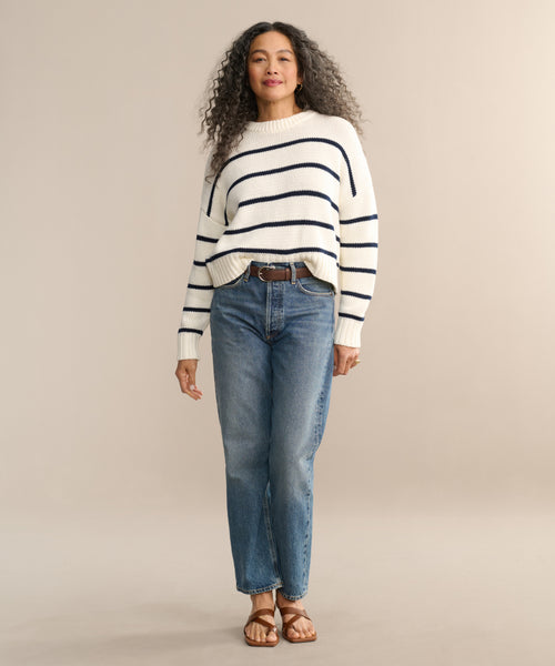 A woman with long curly hair wears a cream sweater with navy stripes, the AGOLDE 90s Jean, and brown sandals. She stands facing forward against a plain beige background.