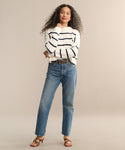 A woman with long curly hair wears a white striped sweater, the 90s Jean from AGOLDE, brown sandals, and a brown belt, standing confidently against a plain beige background.
