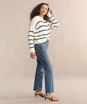 A woman with curly hair stands smiling against a beige background, wearing a white sweater with navy stripes, the 90s Jean by AGOLDE, and brown heeled sandals, with one hand touching her hair.
