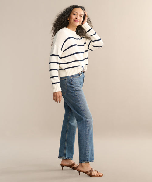 A woman with curly hair stands smiling against a beige background, wearing a white sweater with navy stripes, the 90s Jean by AGOLDE, and brown heeled sandals, with one hand touching her hair.