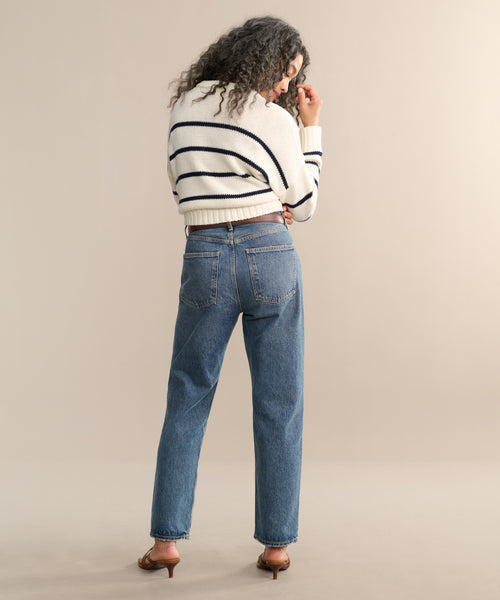 A person with curly hair faces away, wearing a white sweater with black stripes, blue 90s Jean high-waisted AGOLDE jeans made from organic cotton, and brown heeled shoes, set against a plain beige background.