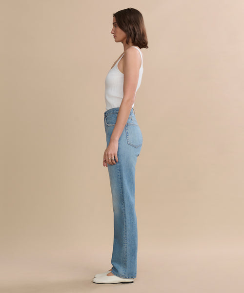 A woman stands in profile against a beige background, wearing a white sleeveless top, the 90s Jean in light blue, and white shoes.