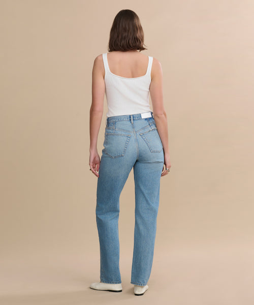 A person with shoulder-length brown hair in a white tank top, 90s Jean light blue high-waisted straight leg jeans, and white shoes stands facing away against a beige background.