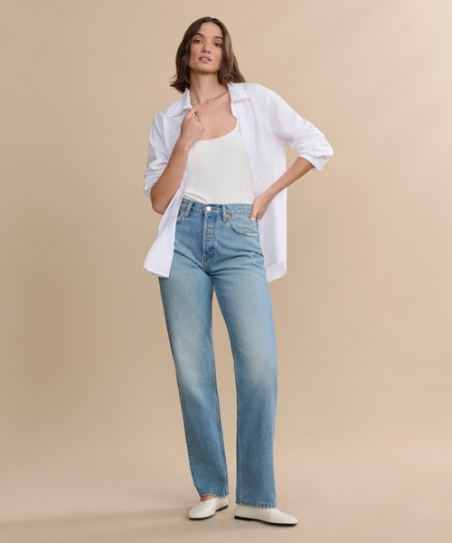 A woman poses against a beige background wearing a white open button-up shirt over a tank top, paired with the 90s Jean in light blue and white shoes. One hand is in her pocket while the other touches her collar.