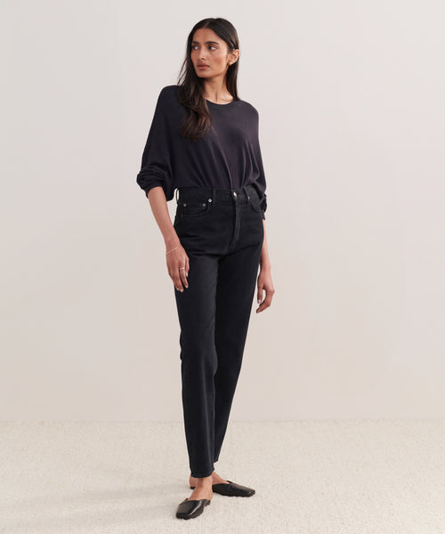 A woman with long dark hair stands against a plain background wearing a loose dark long-sleeve top, black slip-on shoes, and 90s Pinch Waist Long Jeans. She gazes slightly to the side.