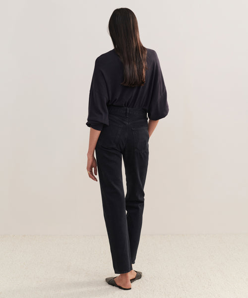 A person with long dark hair, dressed in a loose dark top and 90s Pinch Waist Long Jean, paired with black flats, stands facing away from the camera in a minimalist indoor space with light floors and walls.
