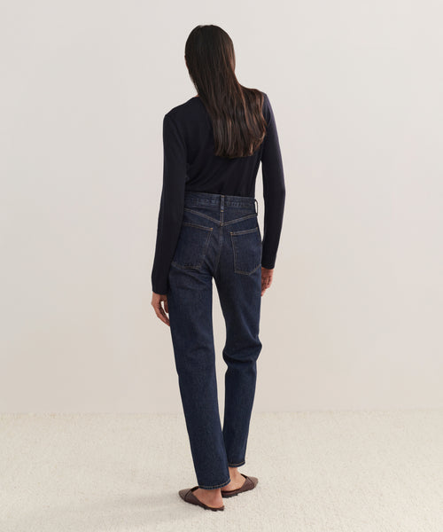 A person with long dark hair, in a black long-sleeve top, 90s Pinch Waist Long Jeans, and brown flats stands facing away from the camera against a plain light background.