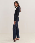 A woman with long dark hair stands sideways against a plain light background, wearing a fitted long-sleeve black top, 90s Pinch Waist Long Jeans in organic cotton, and dark brown flats.