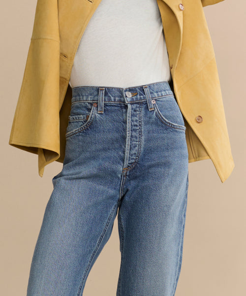 Someone wearing the 90's Pinch Waist Long high-rise straight-leg jeans in comfort stretch denim, styled with a white top and mustard yellow jacket, photographed from waist to upper thighs against a beige background.