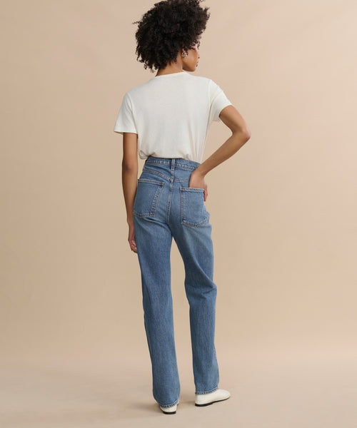 A person stands with their back to the camera, wearing a white short-sleeve t-shirt, 90's Pinch Waist Long high-rise straight-leg comfort stretch jeans, and white shoes against a plain beige background.