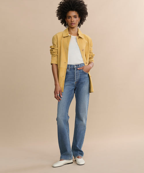 A person with short curly hair wears a yellow jacket over a white shirt, blue 90's Pinch Waist Long jeans in comfort stretch denim, and white shoes, standing against a plain beige background.