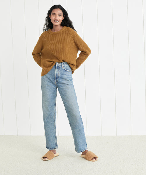 A woman smiles indoors against a white paneled wall, wearing a mustard brown sweater, the 90's Pinch Waist Jean in light blue, and tan fuzzy open-toe slippers.