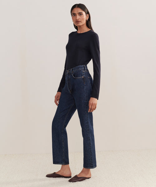 A woman stands sideways against a light background, wearing a fitted black long-sleeve top, 90s Pinch Waist Jean in high-rise blue denim, and brown flat shoes.