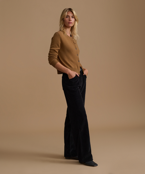 A model wearing a caramel cardigan with black corduroy pants against a beige studio background.