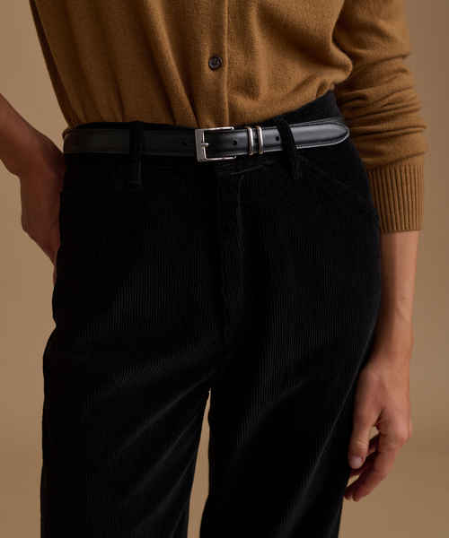 A model wearing a caramel cardigan with black corduroy pants against a beige studio background.