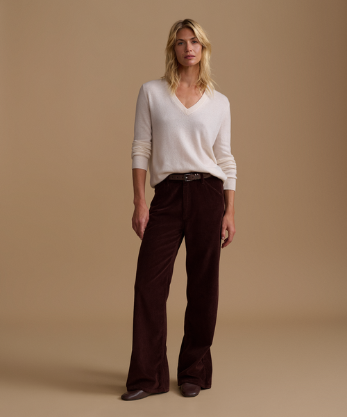 A model wearing an ivory sweater with burgundy corduroy pants against a beige studio background.