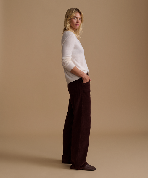 A model wearing an ivory sweater with burgundy corduroy pants against a beige studio background.