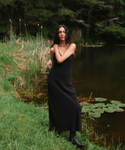 A woman stands on a grassy lakeshore wearing a long black slip-style dress and black boots, with reeds, water, and a dark forest in the background.