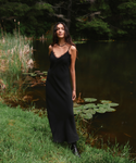 A woman wearing a black lace-trimmed midi slip dress and black ankle boots stands on the grassy bank of a quiet lake, with reeds, lily pads, and a dark forest in the background.