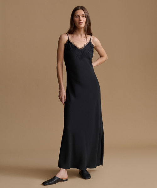 A woman modeling a black slip-style maxi dress with thin straps and lace trim at the neckline, standing against a plain warm beige studio background.