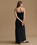 A woman stands with her back to the camera wearing a black satin slip dress with lace trim at the back and thin spaghetti straps, paired with black pointed flats, against a solid beige studio background.