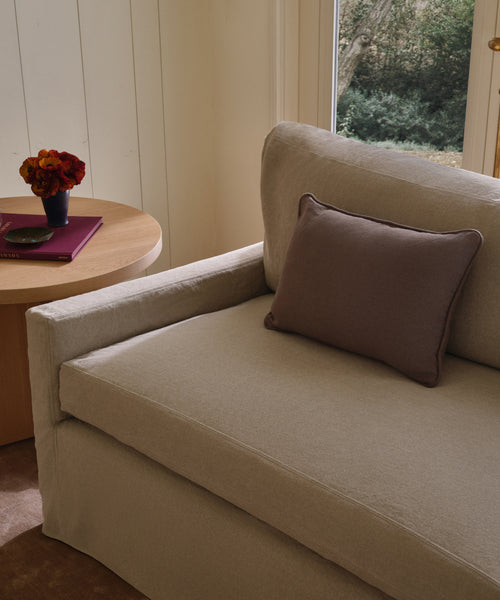 A beige sofa with an Adler Lumbar Pillow sits next to a round wooden side table holding a closed notebook, eyeglasses, and a vase of red and orange flowers. A window behind reveals lush greenery outside.