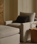 A cozy beige armchair with an Adler Lumbar Pillow rests by a matching ottoman and wooden side table with a vase of white flowers, all set against a cream-paneled wall.