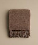 The Alpaca Basketweave Throw, featuring a folded, textured brown design with long fringes at the ends, is displayed on a plain beige background.
