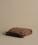 The Alpaca Basketweave Throw, folded with fringed edges, rests on a neutral beige surface against a matching background.
