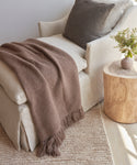 A cozy beige armchair with a brown Alpaca Basketweave Throw made in Peru and a dark cushion sits by a round wooden side table with flowers, atop a light woven rug in a softly lit room.