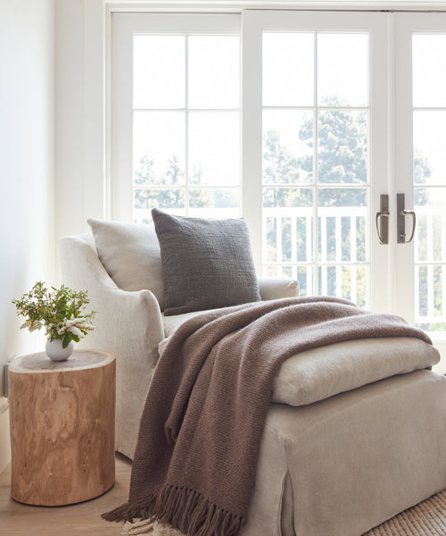 A cozy armchair with a gray pillow and an Alpaca Basketweave Throw sits by large windows with white French doors. A small wooden side table holding a potted plant is beside the chair, all bathed in natural light.