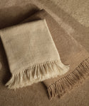 Two Alpaca Basketweave Throws, handwoven in Peru from superfine alpaca—one cream, one light brown with fringed edges—are neatly folded and overlapping on a soft, neutral-toned surface.