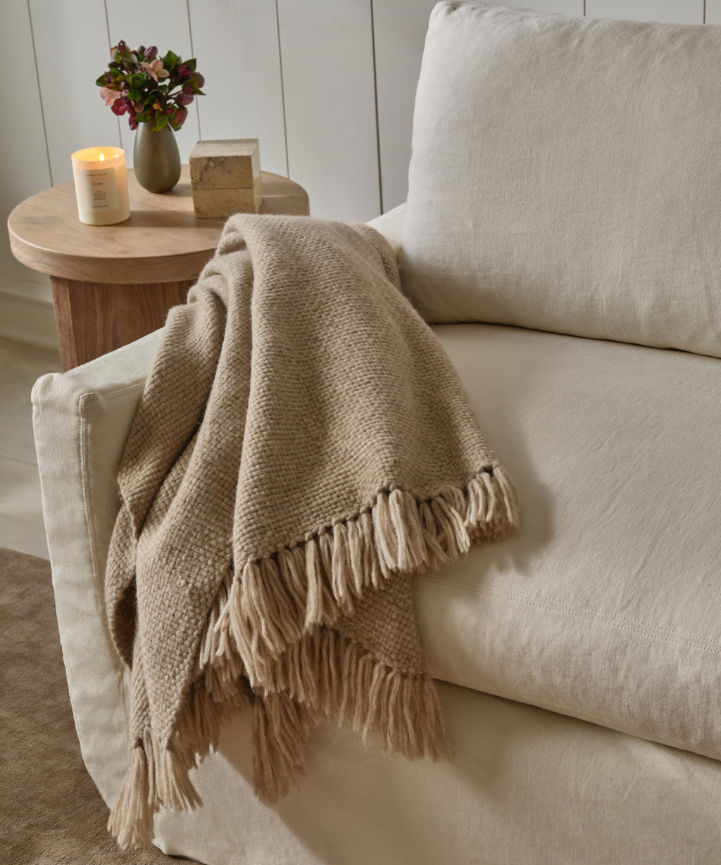 The Alpaca Basketweave Throw, a beige handwoven Peruvian alpaca blanket with fringe, is draped over a light sofa. Nearby, a wooden side table with a lit candle and small vase adds to the cozy scene.