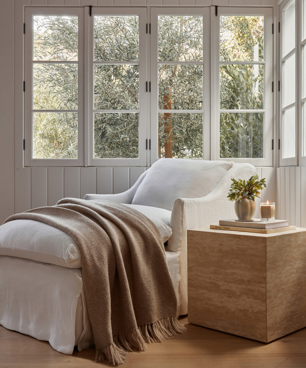 A cozy white armchair with an Alpaca Basketweave Throw and cushion sits by large windows; nearby, a travertine side table holds a vase of greenery and a lit candle. Sunlight streams through the trees outside.