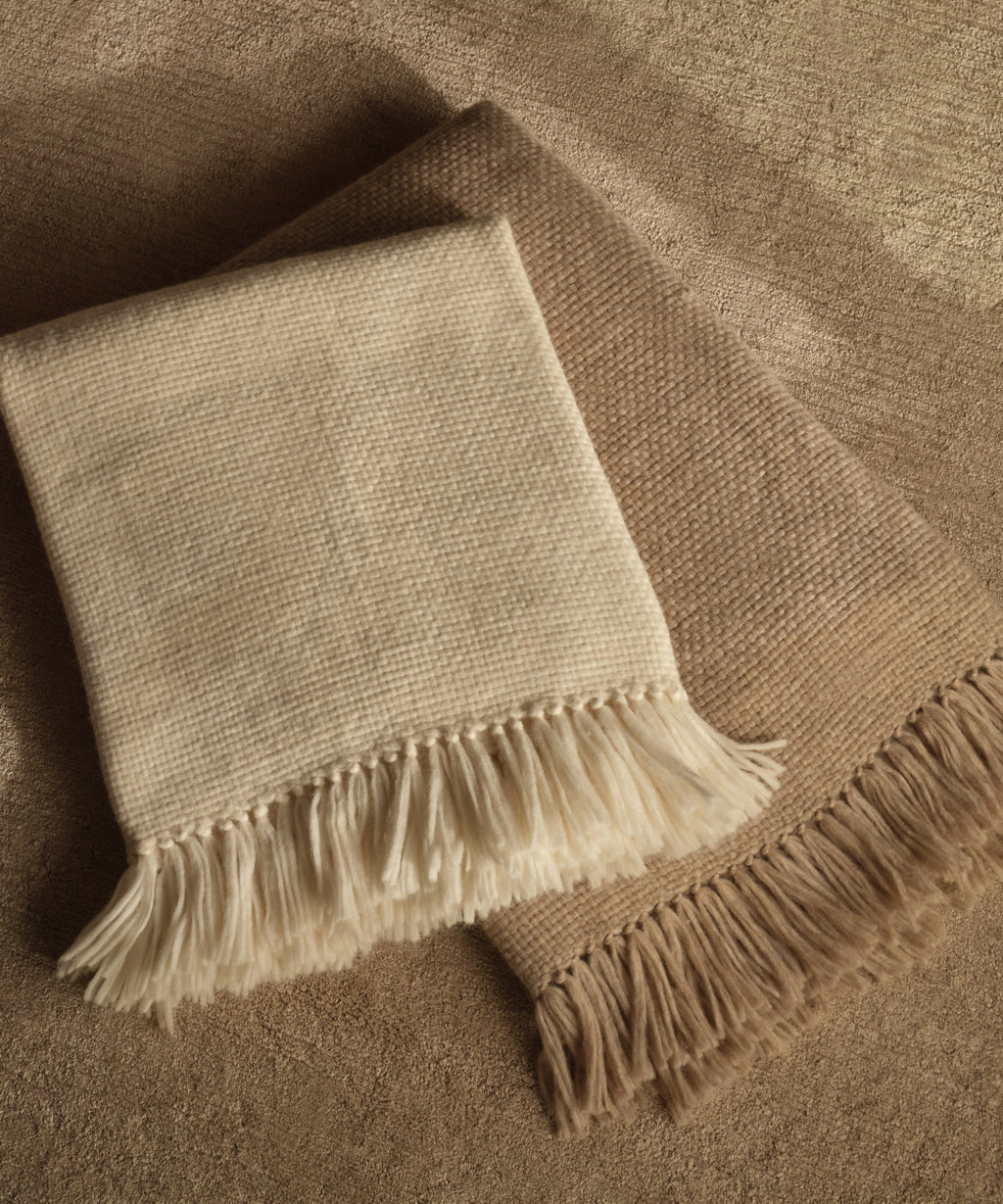 Two Alpaca Basketweave Throws—one cream and one light brown—are neatly folded and overlapping on a textured beige surface. Handwoven in Peru from superfine alpaca, they offer exceptional softness and warmth.